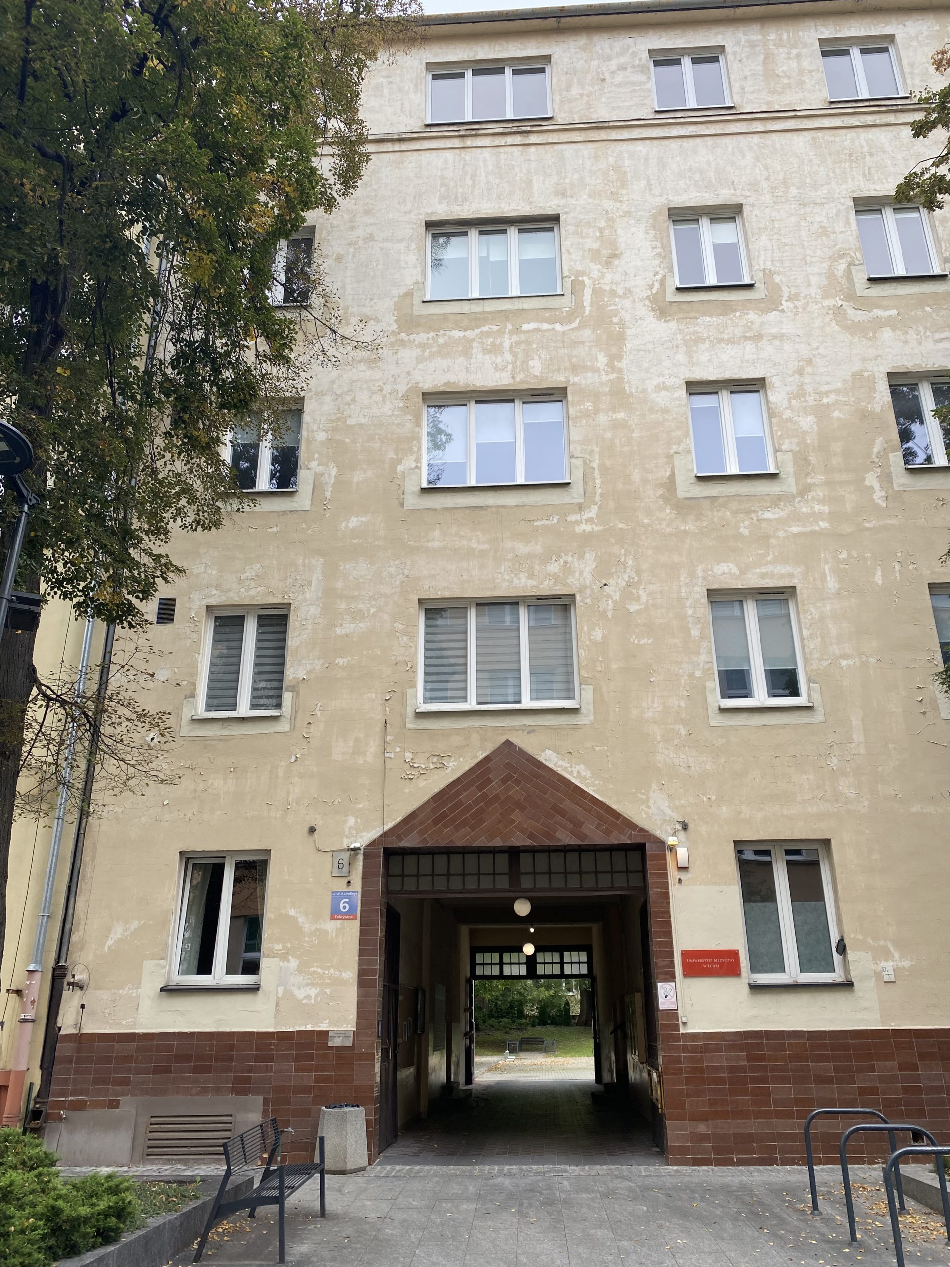 Elewacja budynku z przejściem. Budynek o jasnej, kremowej elewacji z widocznymi śladami zużycia. Na parterze centralnie umieszczone przejście bramowe z brązową podmurówką i daszkiem w kształcie trójkąta. Nad przejściem i po bokach rzędy prostokątnych okien. Po lewej stronie widoczne drzewa, po prawej stojaki na rowery.