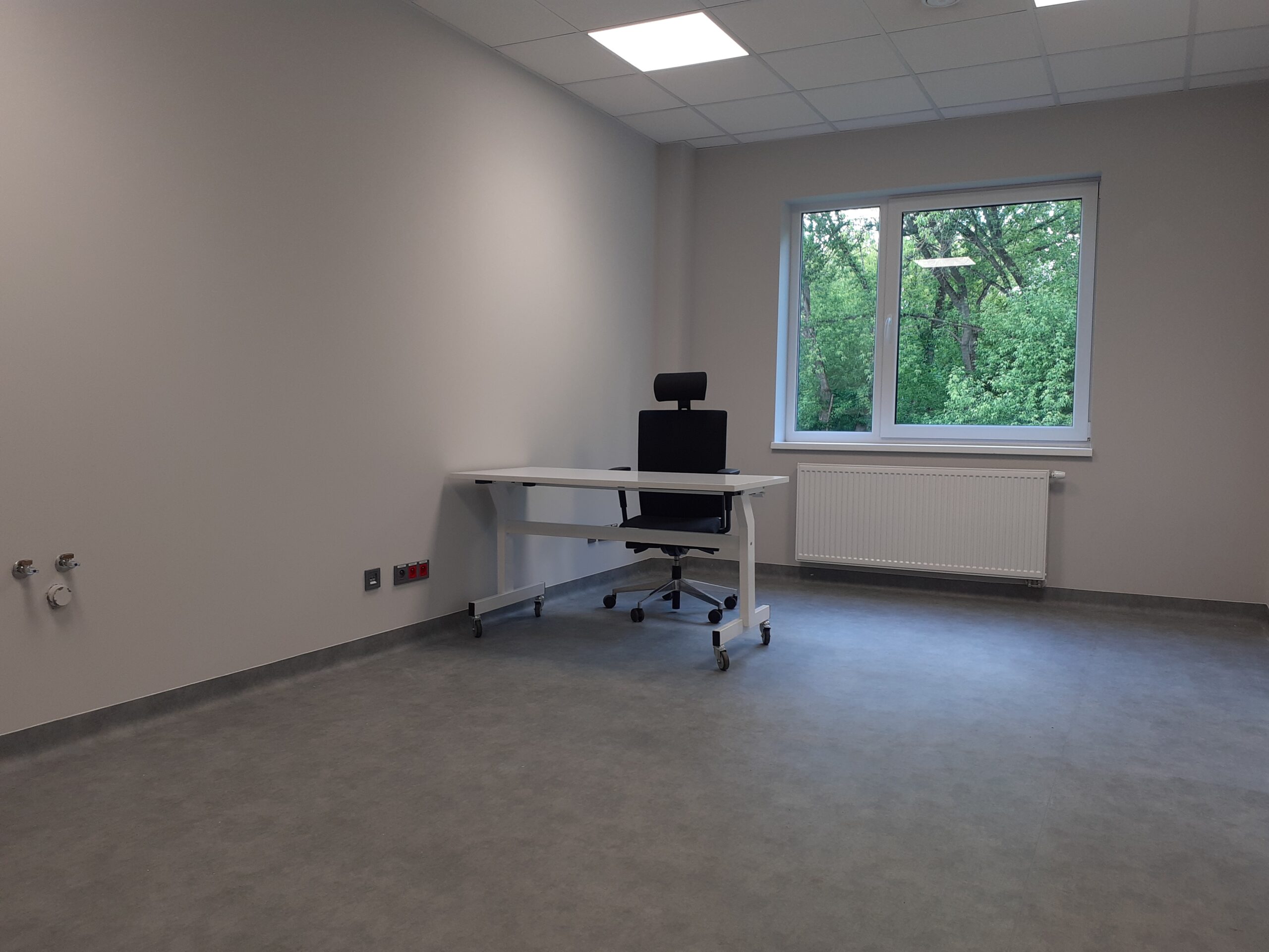 Po lewej stronie na dłuższej ścianie są widoczne nad podłogą oddalone od siebie dwa punkty z gniazdkami. Po lewej stronie na dłuższej ścianie widoczne jest biurko i krzesło