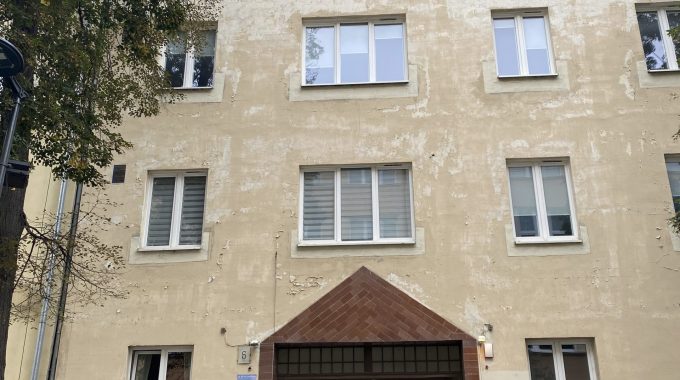 Elewacja budynku z przejściem. Budynek o jasnej, kremowej elewacji z widocznymi śladami zużycia. Na parterze centralnie umieszczone przejście bramowe z brązową podmurówką i daszkiem w kształcie trójkąta. Nad przejściem i po bokach rzędy prostokątnych okien. Po lewej stronie widoczne drzewa, po prawej stojaki na rowery.