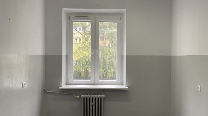 Wnętrze pustego pokoju z oknem. Prostokątne pomieszczenie z białymi ścianami i ciemnoszarą wykładziną na podłodze. Na krótszej ścianie znajduje się okno z białą ramą, pod nim grzejnik żeliwny. Na suficie podłużna lampa świetlówkowa. Pomieszczenie jest puste, bez mebli.