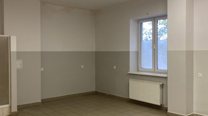 Pomieszczenie z płytkami na podłodze. Prostokątne pomieszczenie z jasnoszarymi ścianami i beżową podłogą z dużych płytek. Na krótszej ścianie okno z białą ramą, pod nim grzejnik panelowy. Na suficie kilka prostokątnych lamp LED.