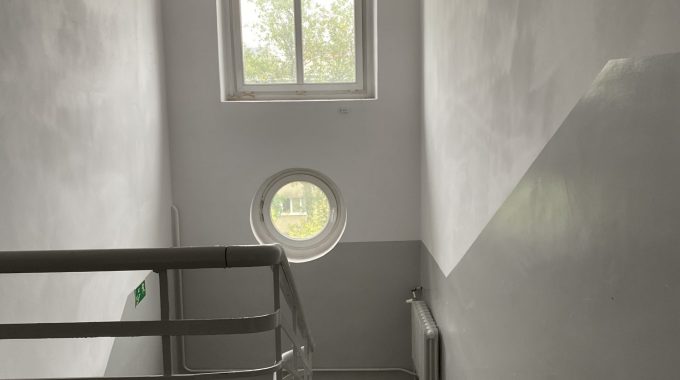 Klatka schodowa z oknem okrągłym. Wnętrze klatki schodowej z betonowymi schodami i białą metalową balustradą. Na ścianie okrągłe okno oraz prostokątne okno powyżej. Ściany i sufit pomalowane na biało i jasnoszaro, podłoga jasnoszara.