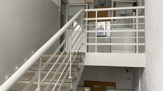 Ostatnie piętro z klatką schodową i tablicami. Widok z półpiętra na klatkę schodową z białą metalową balustradą. W tle ściana z tablicami informacyjnymi i gablotami. Podłoga jasnoszara, ściany białe, sufit z prostokątnymi lampami.