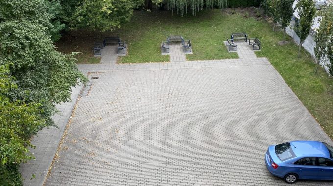 Parking z samochodami i zielenią. Utwardzony parking z kostki brukowej, na którym stoją dwa samochody: czerwony i niebieski. Po prawej stronie pas zieleni z drzewami i krzewami, na wprost ławki. W tle widoczne budynki mieszkalne.